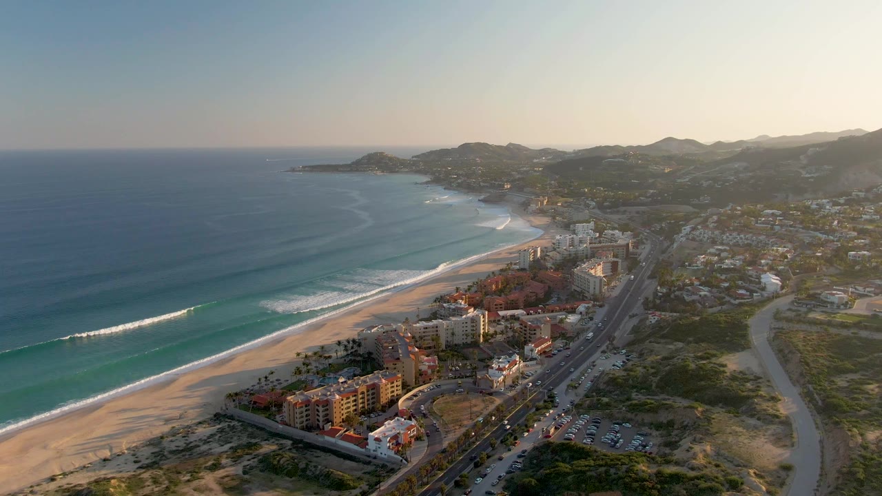 Aerial frame over San Jose del Cabo with blue ocean and surf on the left, beachside buildings and a parallel road below, hills in the distance, and no visible people.