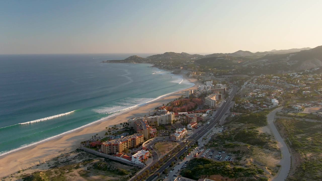 Drone view along a sandy coastline with surf, road traffic, buildings, and hills.