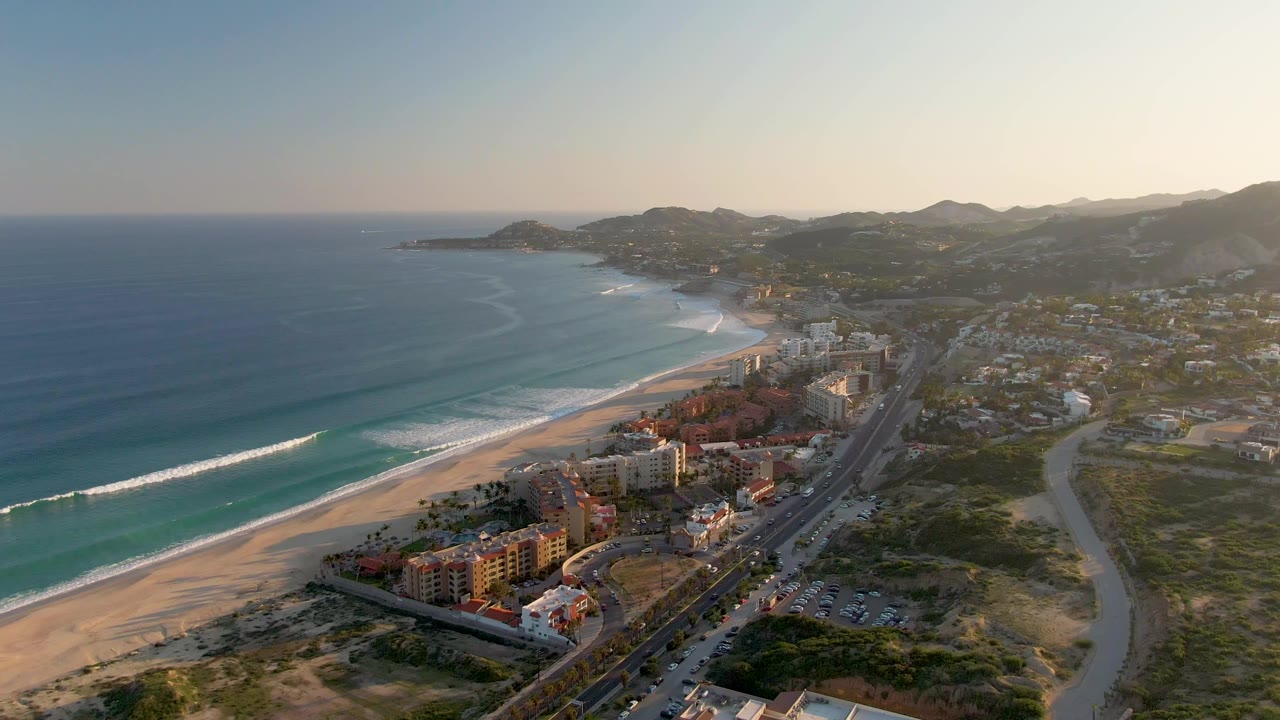 A high aerial frame of San Jose del Cabo shows the beach curving toward the hills, with turquoise water, roadside buildings, moving vehicles, and no visible people.
