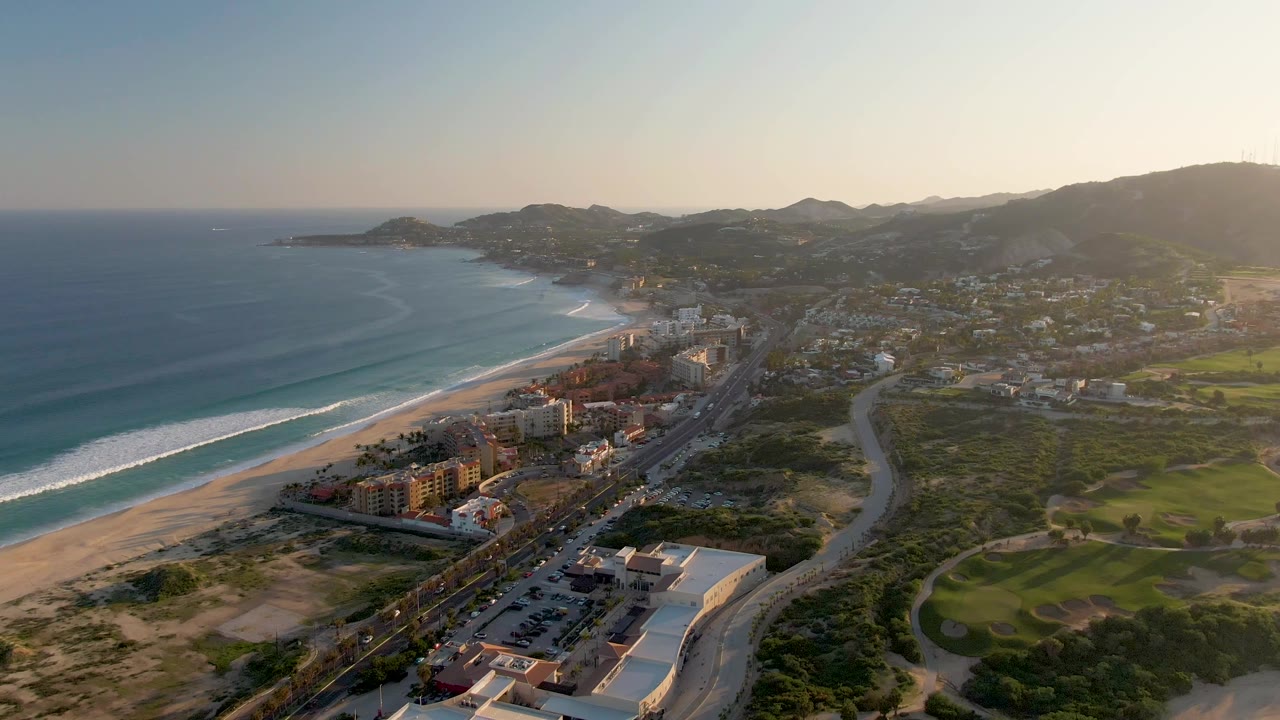 Aerial view over San Jose del Cabo with blue ocean and surf at left, a sandy beach below, a road and buildings through the center, green fields to the right, and no visible people.