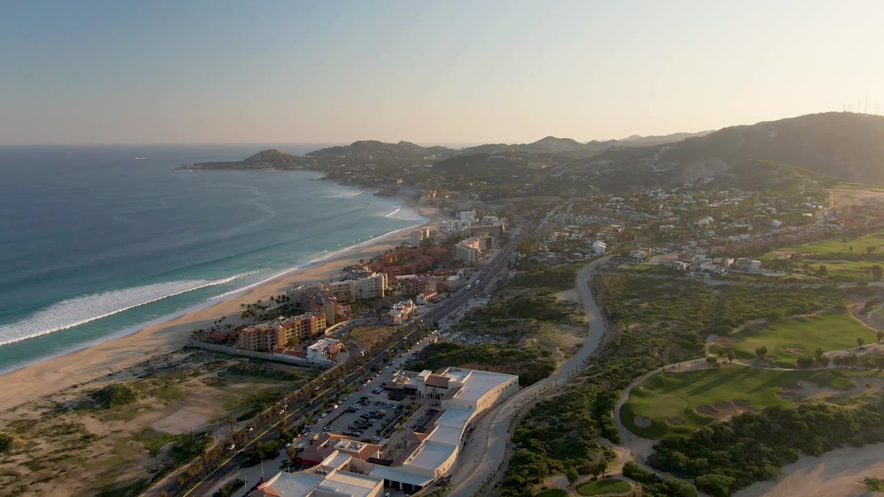 Wide San Jose del Cabo aerial showing the ocean on the left, white surf bands along the beach, shoreline buildings and a road in the middle distance, hills beyond, and no visible people.