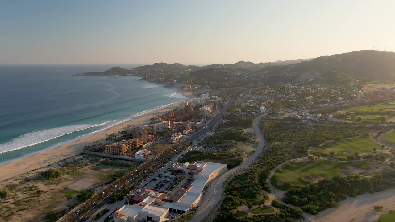 San Jose del Cabo coastline from above, with the beach and ocean at left, clusters of buildings near the shore, a winding road crossing the right side, green terrain, hills, and no visible people.