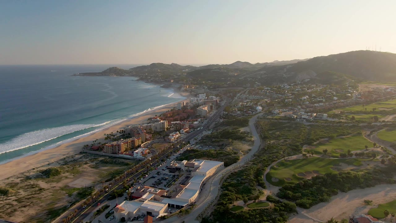 Aerial view over San Jose del Cabo with surf rolling toward the sandy beach, a line of buildings beside the coast, a road curving inland, green fields on the right, distant hills, and no visible people.