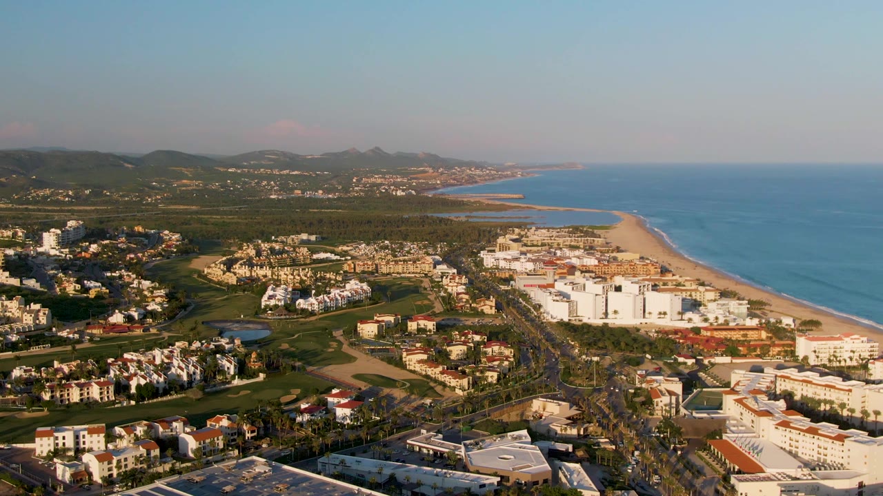 Opening aerial frame over San Jose Del Cabo, Mexico, showing resort buildings, a road corridor, green golf course areas, sandy beach, blue ocean, distant hills, and no visible people.