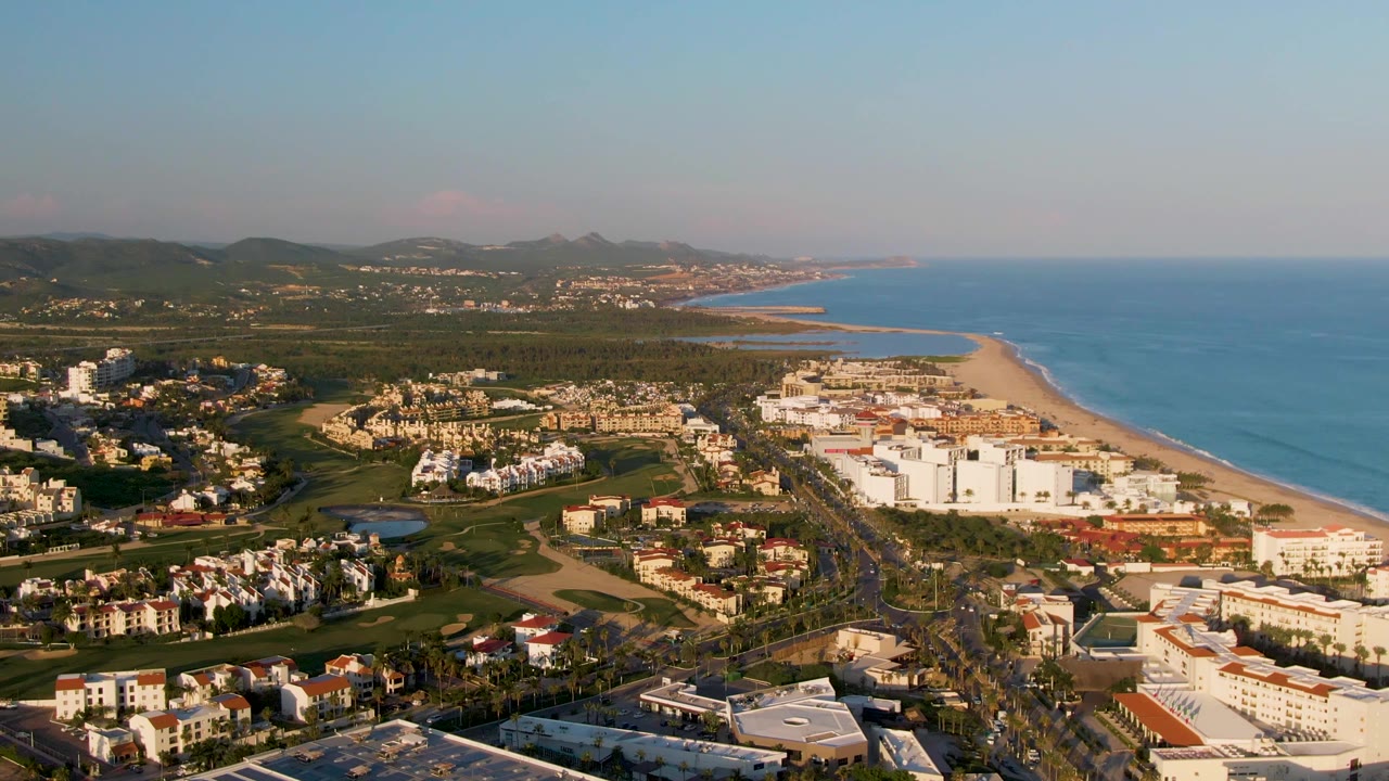One second into the San Jose Del Cabo aerial, the view holds on white resort blocks, villas, a road running toward the beach district, blue water, low hills, and no visible people.