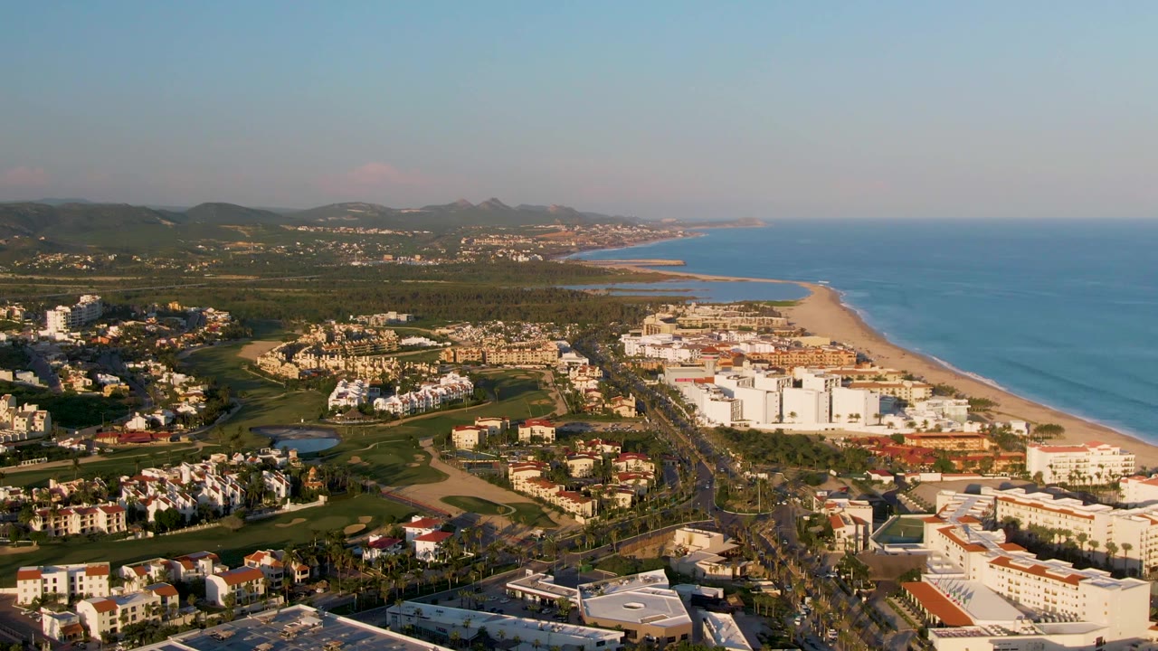 Aerial view across coastal resort buildings toward a long beach, ocean, and inland hills.