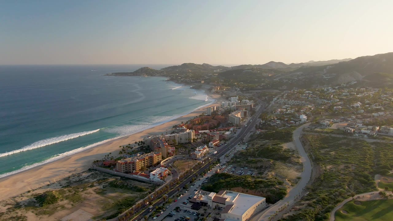 Aerial opening frame over San Jose del Cabo with blue ocean and surf at left, sandy beach below, coastal highway and buildings through the center, hills in the distance, and no visible people.