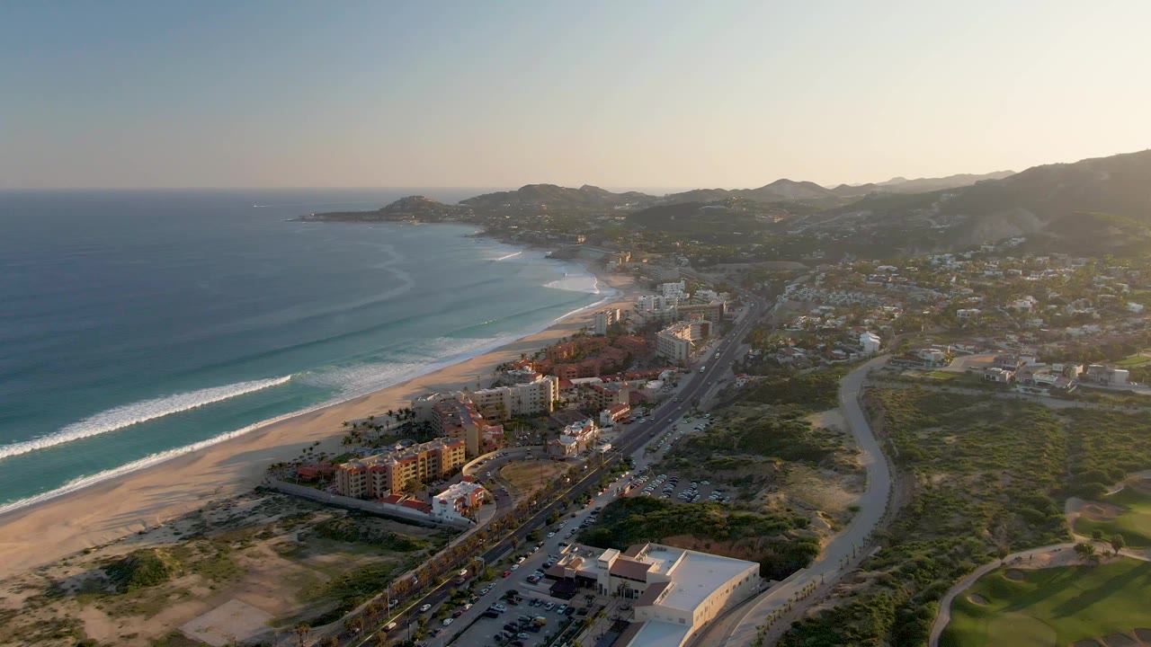 San Jose del Cabo aerial frame with a longer stretch of white surf visible along the beach, highway traffic beside shoreline buildings, green open land to the right, and no visible people.