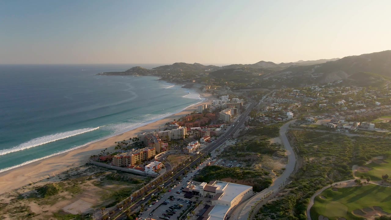 Wide San Jose del Cabo coastline view with ocean filling the left side, a bright surf band on the beach, a road corridor and clustered buildings in the middle, green course-like terrain at right, and no visible people.