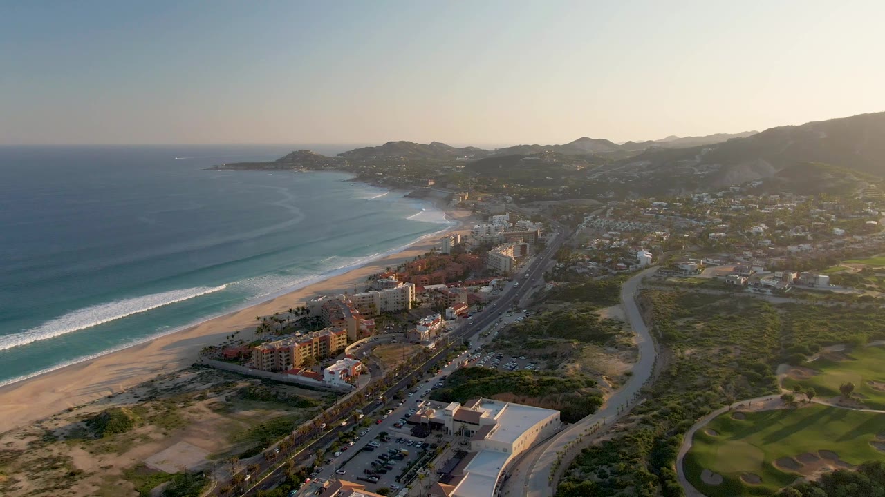 San Jose del Cabo aerial frame with the beach and surf sweeping diagonally from lower left, compact buildings beside the highway, green terrain and hills to the right, and no visible people.