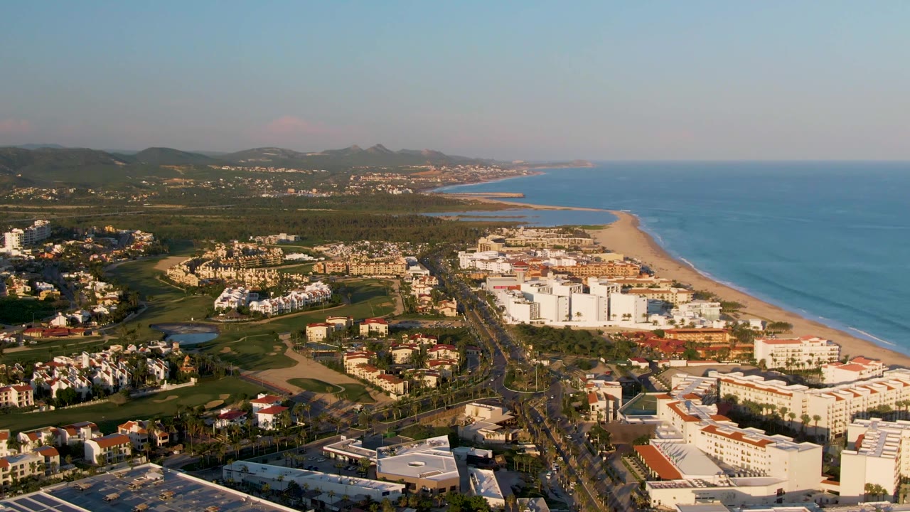 Opening aerial frame over San Jose del Cabo showing resort and residential buildings beside a sandy beach, blue ocean, a central road with cars, green terrain, distant mountains, and no visible people.