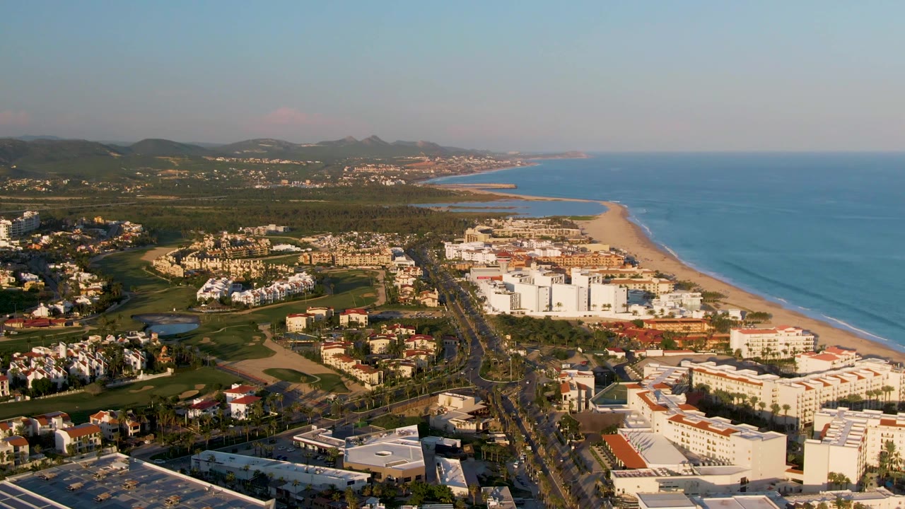 One second into the San Jose del Cabo aerial, the coastline remains broad in frame with white resort blocks, the beach running along the ocean, cars on the central road, green land, and no visible people.