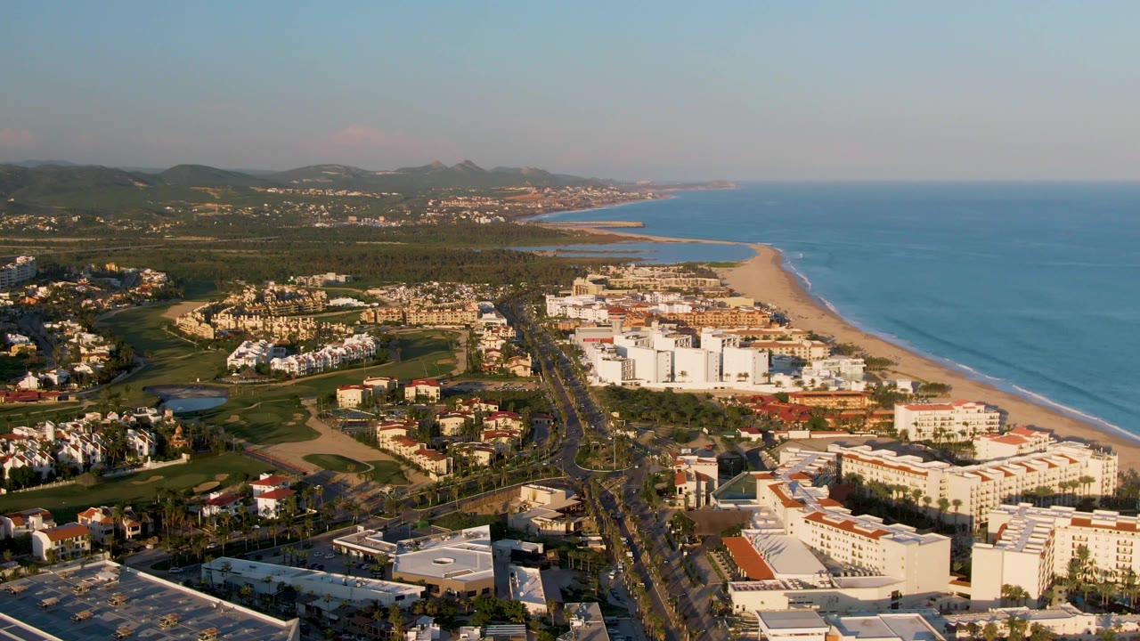 At 0:02, the San Jose del Cabo drone view shows more of the beach and shoreline to the right, with the road and resort buildings below, green inland areas, distant hills, and no visible people.