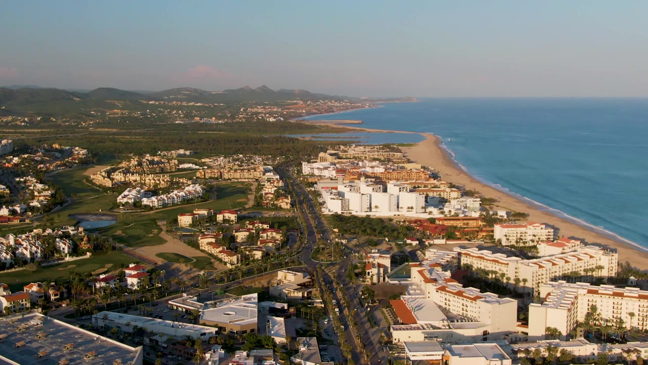 Drone view along beach and resort area with road, ocean, greenery, and distant mountains.