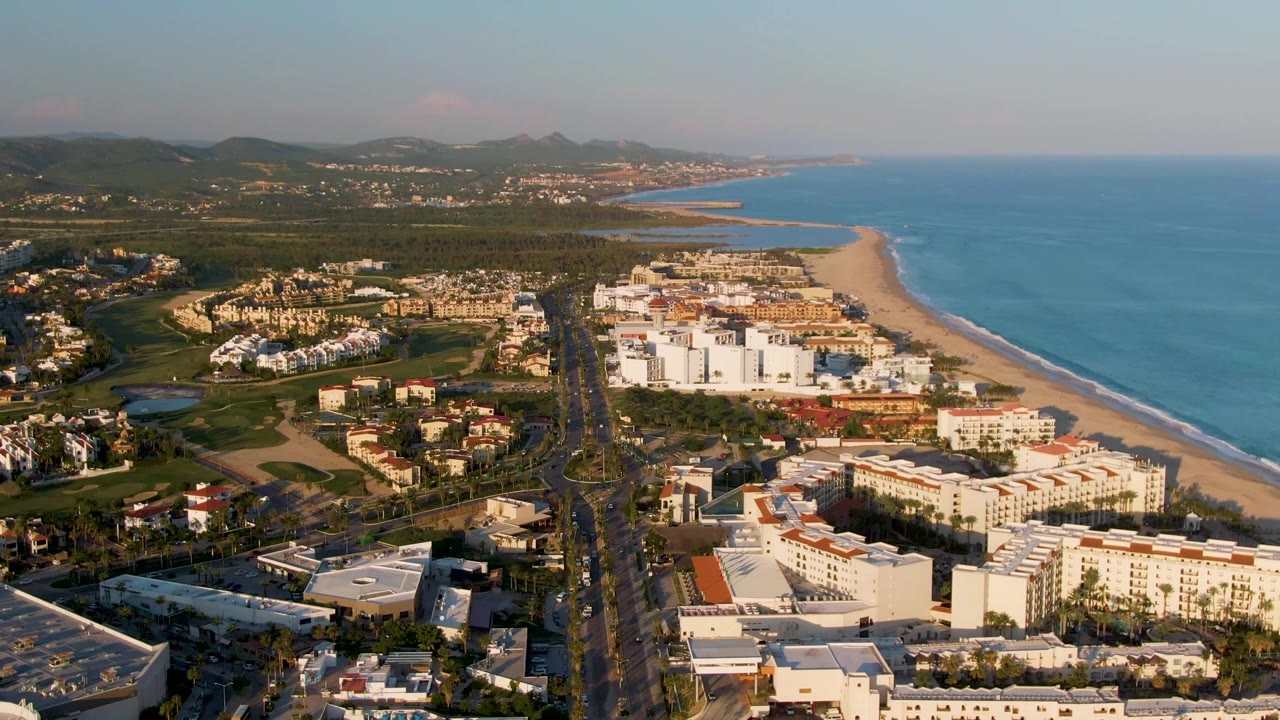 Aerial frame over San Jose Del Cabo showing a coastal highway running toward white beachfront buildings, a sandy beach and blue ocean on the right, green areas and hills beyond, with no visible people.