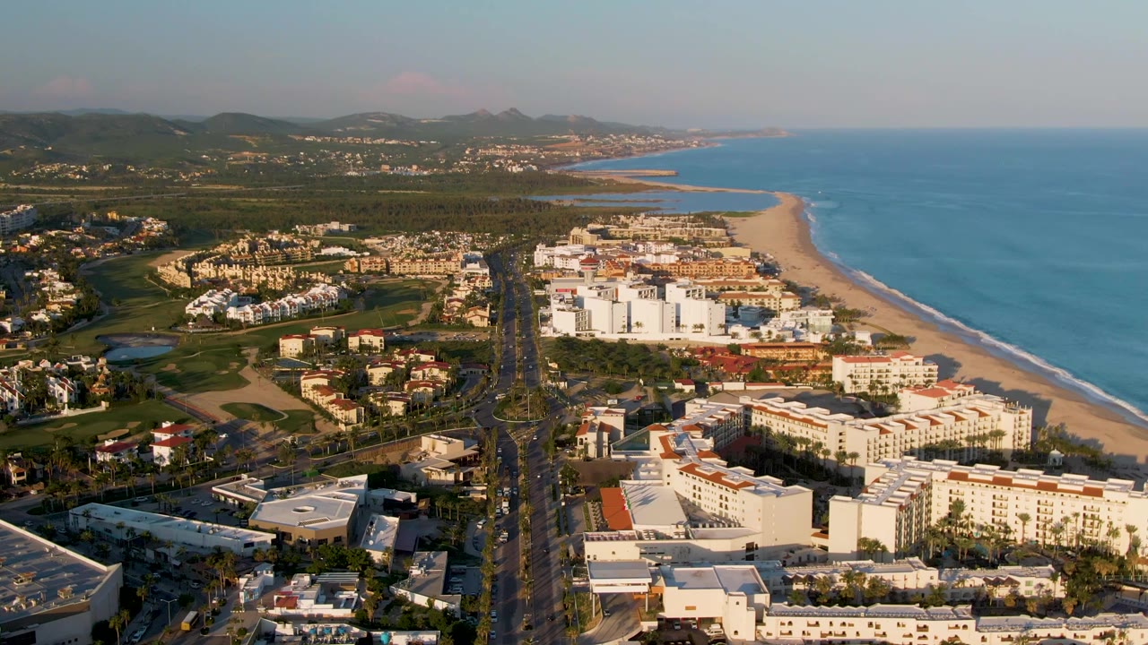 Aerial frame in San Jose Del Cabo with the highway centered below, compact white and tan buildings near the beach, ocean to the right, green land to the left, and no visible people.