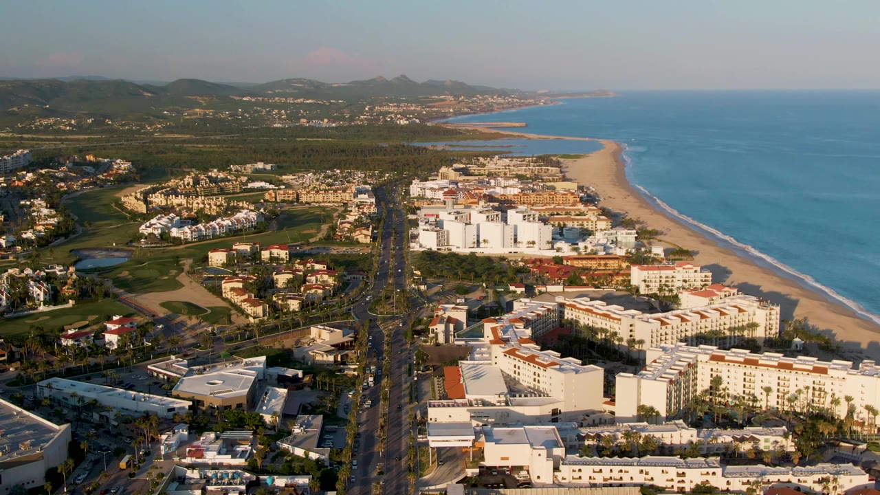 Aerial frame over San Jose Del Cabo with more of the beachfront district visible, including pale buildings, a long beach strip, blue water, inland hills, and no visible people.