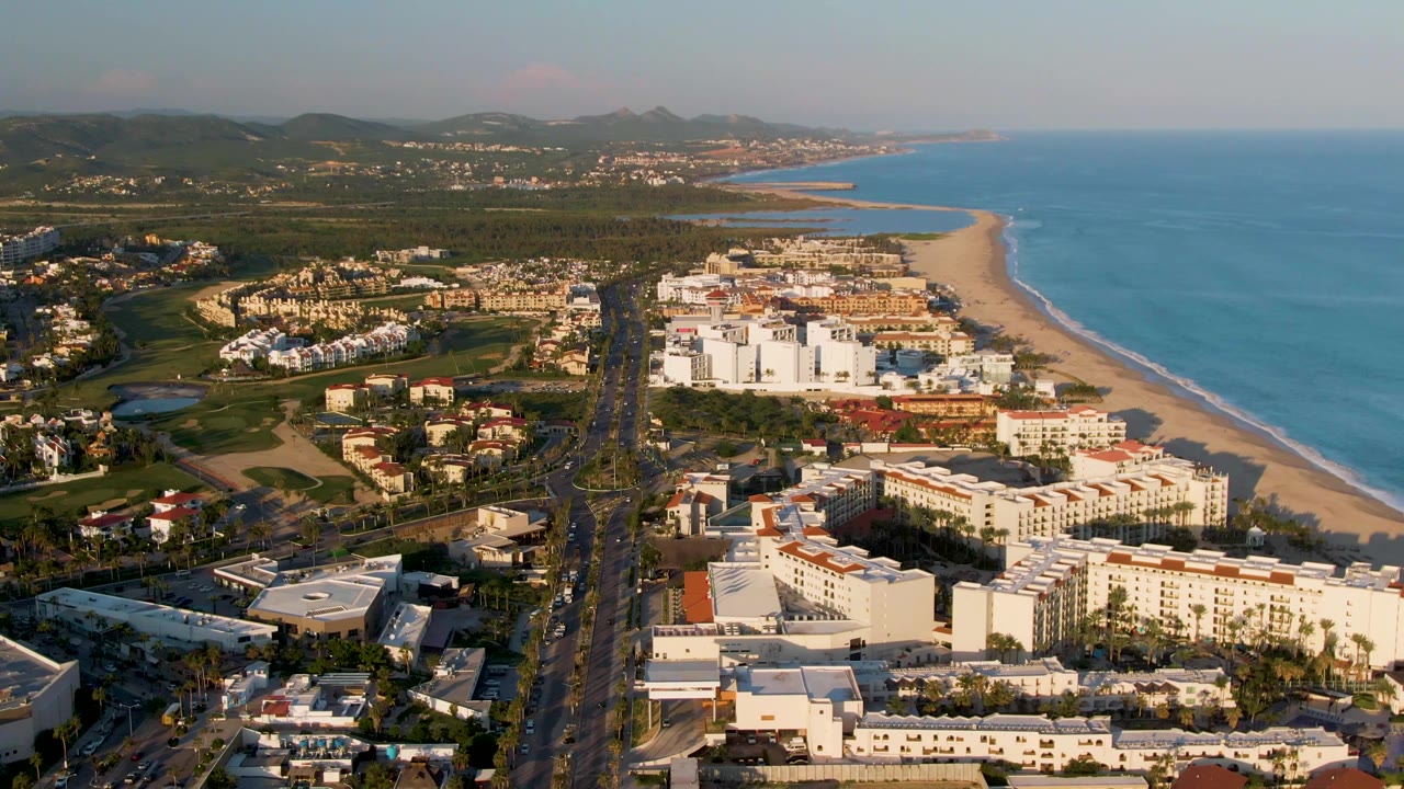 Coastal aerial with highway traffic, resort-like buildings, beach, ocean, green land, and distant hills.