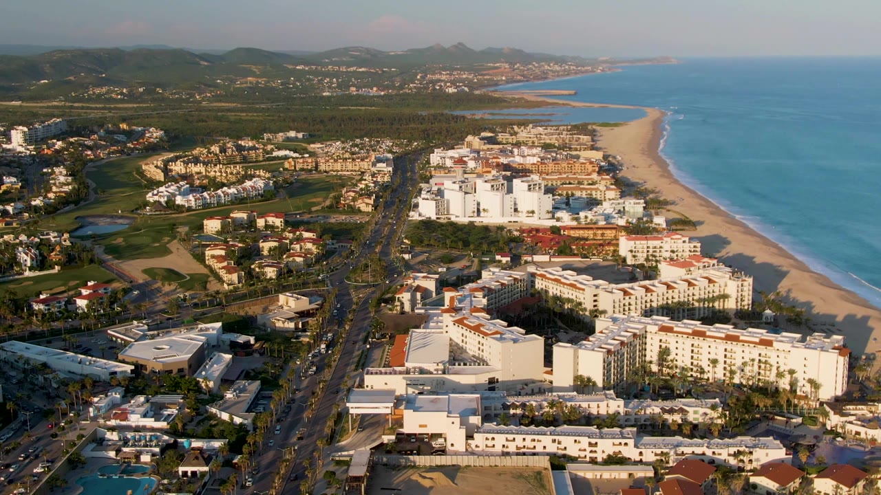 Aerial view of beachside buildings, road, sandy coast, ocean, and green hills.