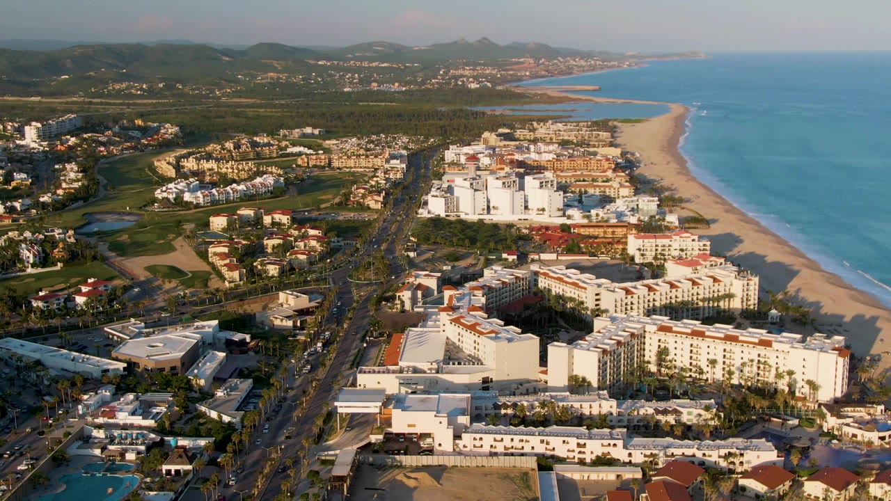 High aerial frame of San Jose del Cabo showing clustered white resort-style buildings near the beach, the ocean running along the right side, a road through the center, and no visible people.
