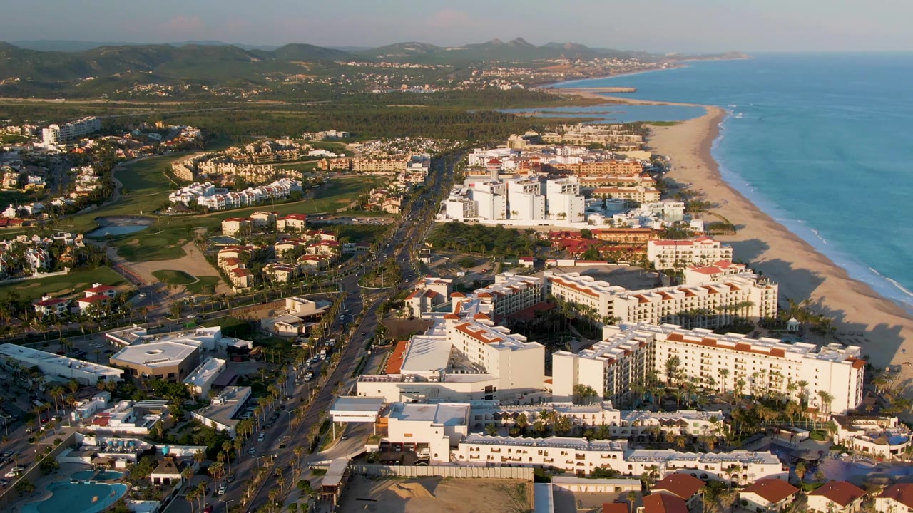Aerial view in San Jose del Cabo with sunlit hotel blocks in the foreground, a multi-lane road carrying vehicles, a long sandy beach, blue water, distant hills, and no visible people.