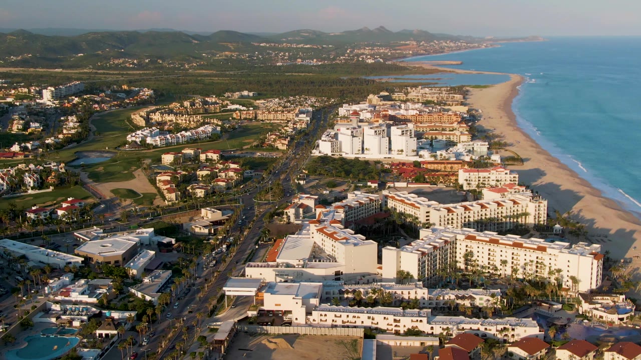Drone-height view of San Jose del Cabo with the road and vehicles leading through white beachfront buildings, green inland terrain to the left, the sandy beach and ocean to the right, and no visible people.