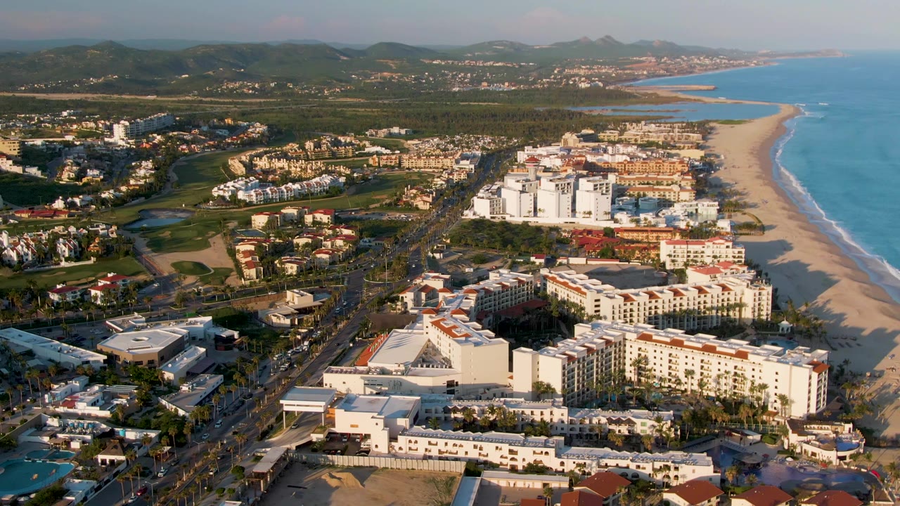 Aerial frame over San Jose del Cabo with beachfront hotel blocks, a road with vehicles, sandy beach, blue ocean, inland greenery, distant hills, and no visible people.
