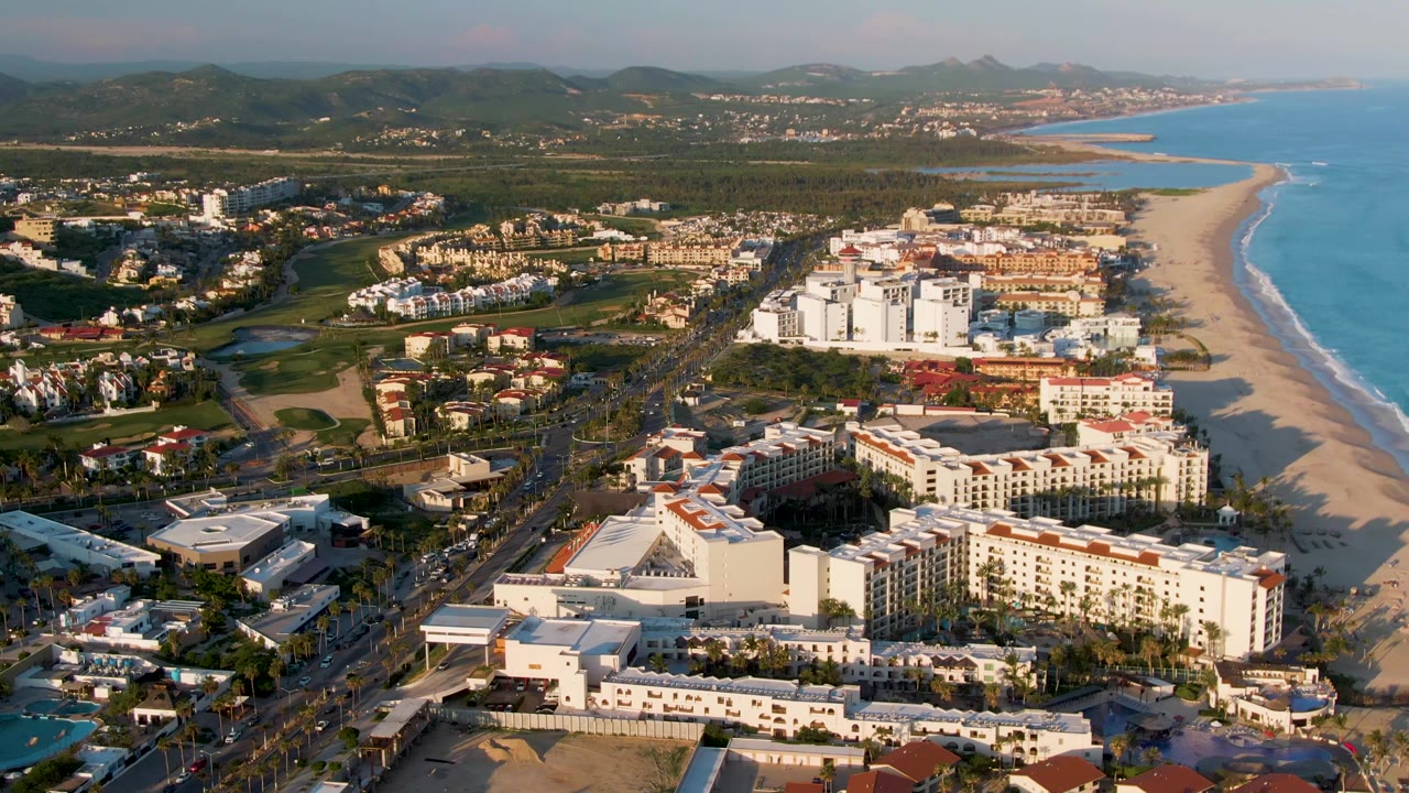 Wide aerial frame in San Jose del Cabo showing white beachfront buildings, palm-lined blocks, road traffic, a long sandy beach, ocean surf, and no visible people.