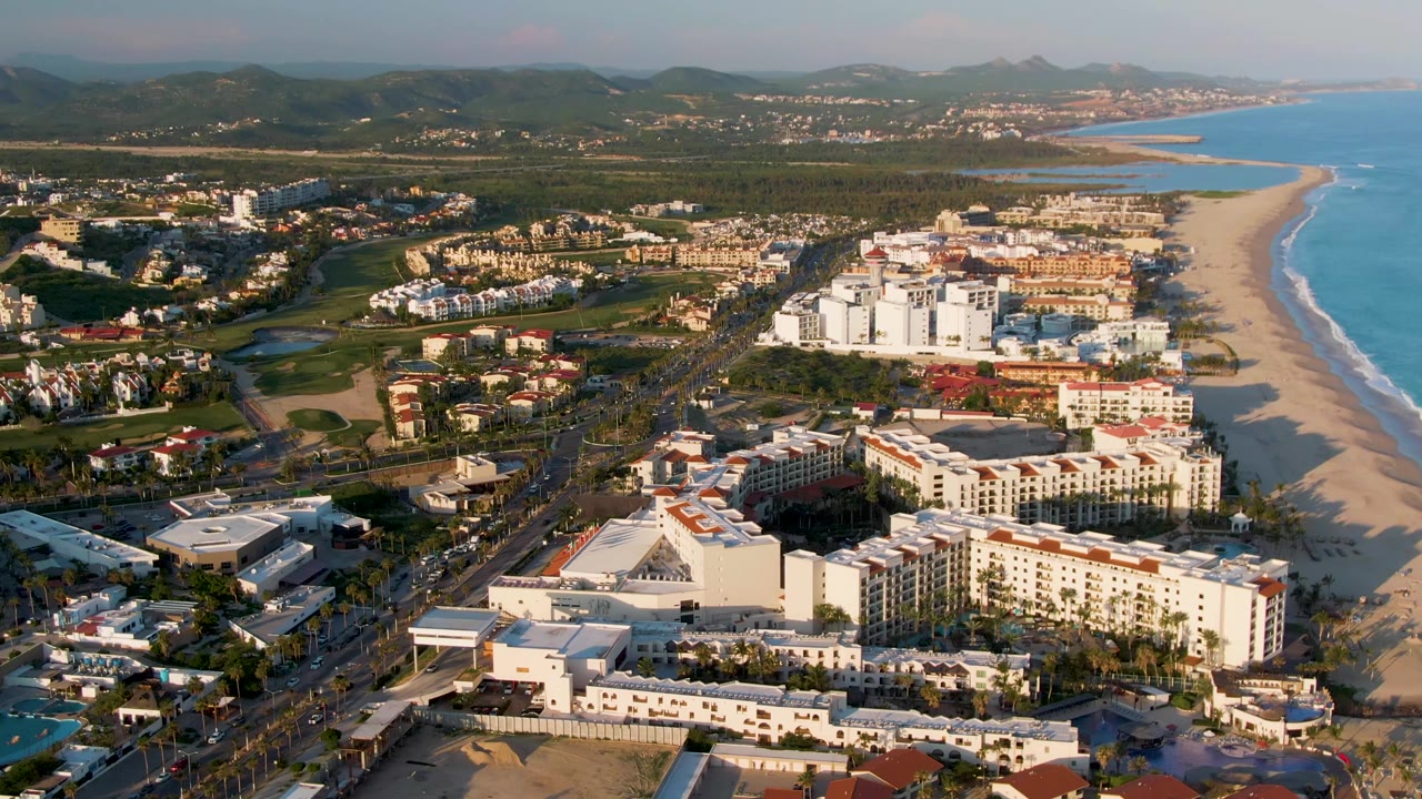 San Jose del Cabo aerial frame with foreground rooftops, beachfront development, small vehicles on the main road, a pale sand beach, blue water, hazy hills, and no visible people.