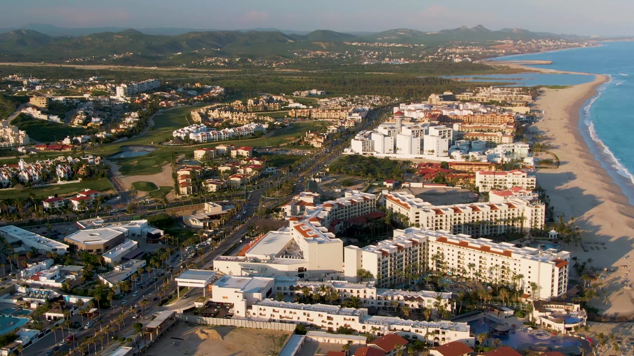 Aerial coastal view with resort buildings, road traffic, beach, ocean, greenery, and hills.