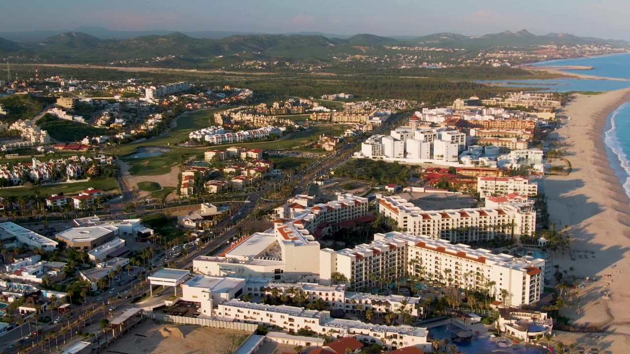 Aerial view over San Jose Del Cabo resort buildings with a wide beach and ocean to the right, roads and greenery through the middle, mountains on the horizon, and no visible people.