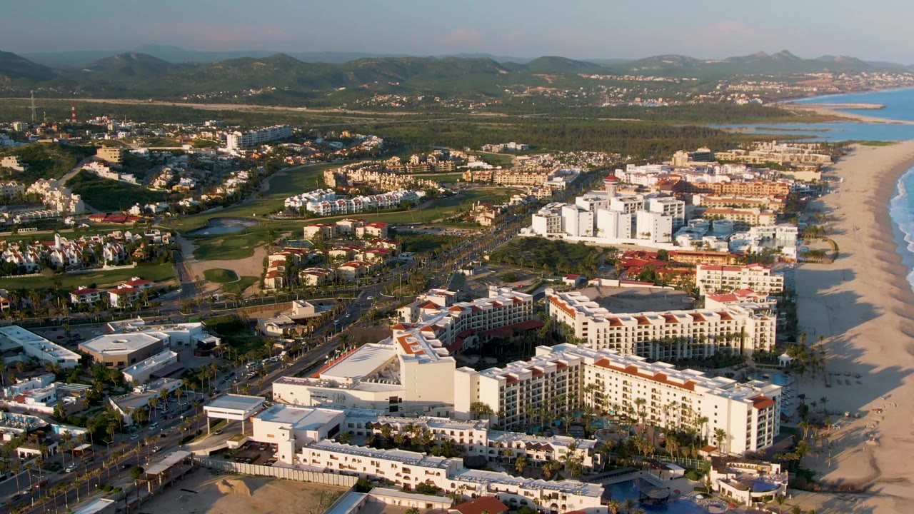 High aerial frame over San Jose Del Cabo showing white and tan hotel blocks near the beach, ocean water at frame right, inland roads, green areas, distant hills, and no visible people.