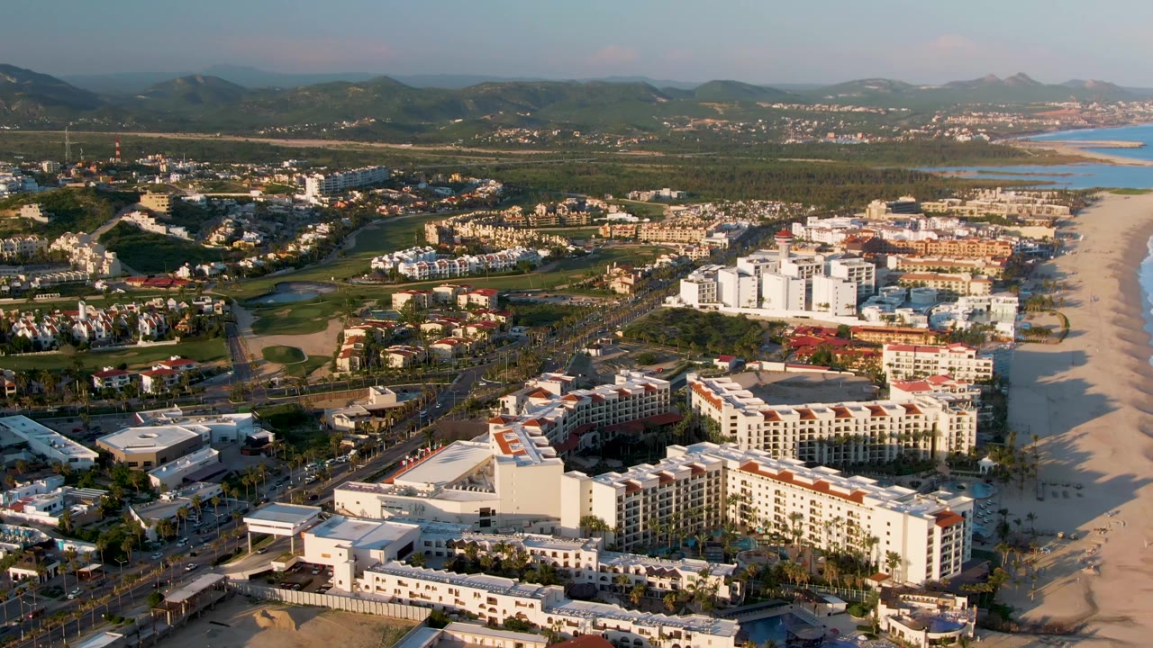 Drone-style aerial over San Jose Del Cabo with a large resort complex in the lower frame, beach and surf line to the right, town and greenery inland, mountains beyond, and no visible people.