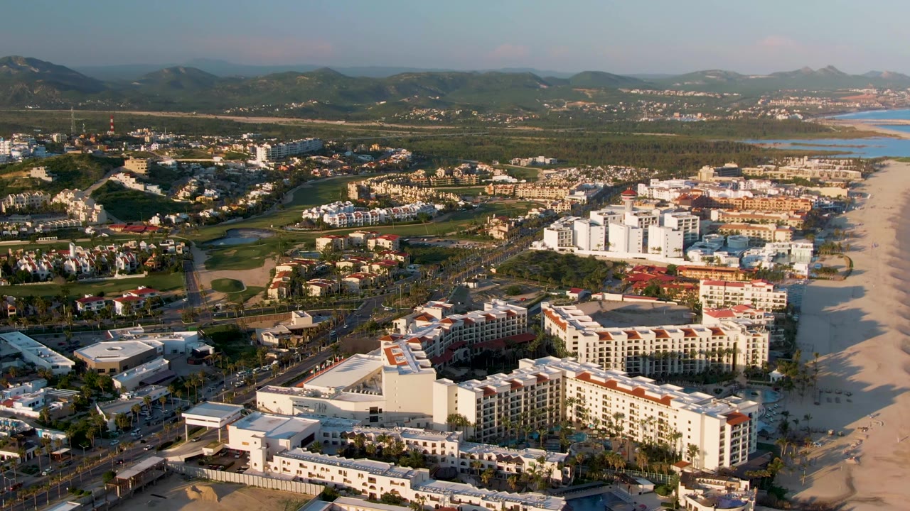 Wide aerial view of coastal hotels, beach, ocean, roadways, greenery, and mountain horizon.