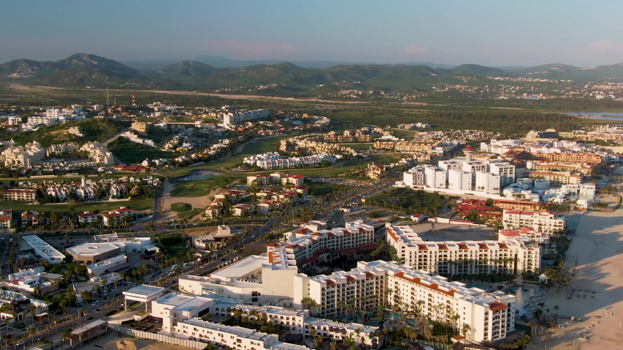 Aerial view over San Jose Del Cabo with beachfront sand at the lower edge, clustered buildings, roads with small vehicles, green areas, distant mountains, and no visible people.