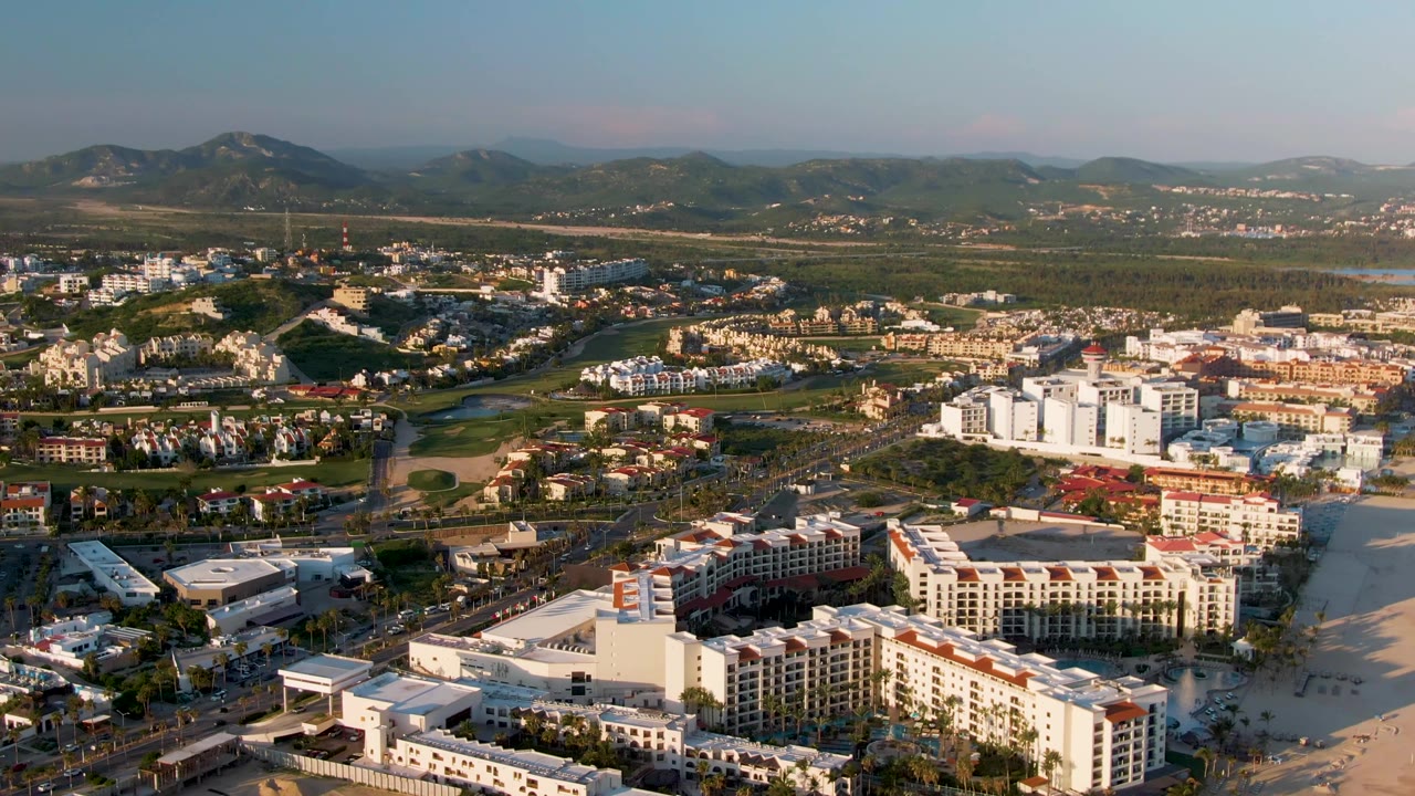 Wide aerial over San Jose Del Cabo showing resort-scale buildings beside the beach, a road network with tiny cars, green patches inland, distant hills, warm light, and no visible people.