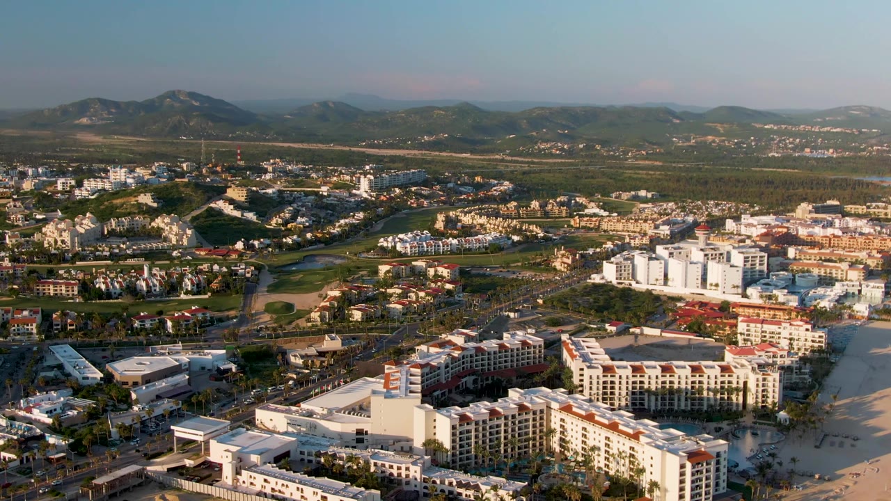 A San Jose Del Cabo aerial frame with a large beachfront complex in the lower area, streets and moving-size vehicle dots through the center, tree-lined green zones, mountains beyond, and no visible people.