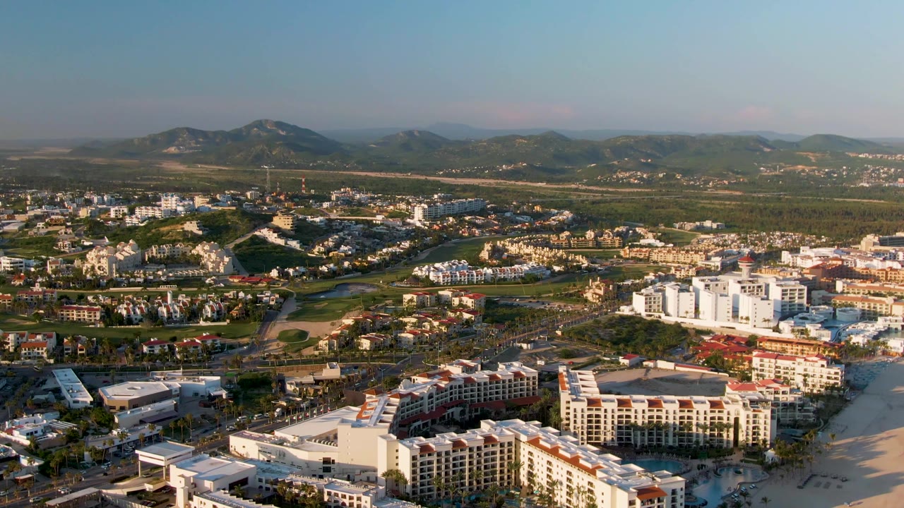 Opening aerial frame over San Jose Del Cabo resort buildings and beach, with town blocks, greenery, hazy mountains, and no visible people.