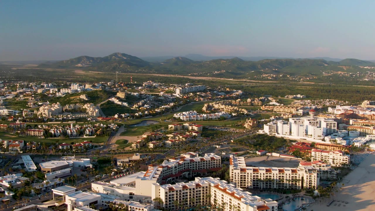 Second aerial frame in San Jose Del Cabo showing resort roofs near the sand, scattered town development, green hills, distant mountains, and no visible people.
