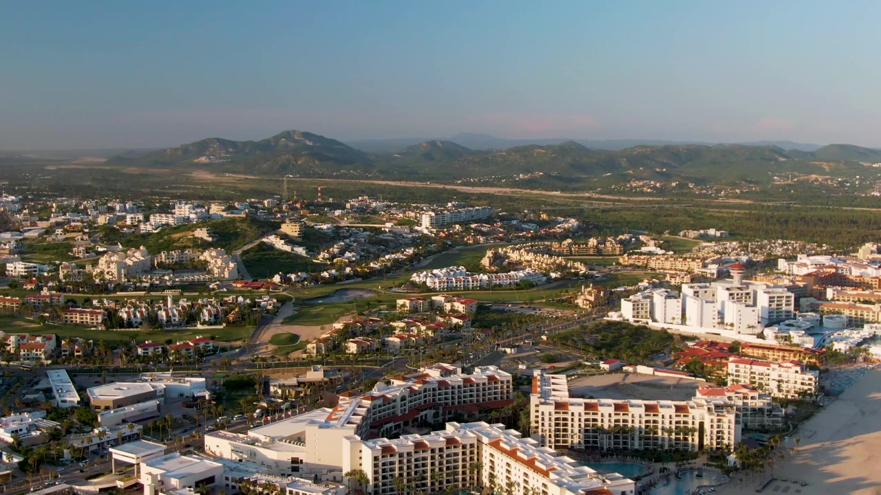 Mid-clip aerial frame of San Jose Del Cabo with bright resort buildings beside pale sand, low-rise urban blocks, a broad green plain, mountain ridges, and no visible people.