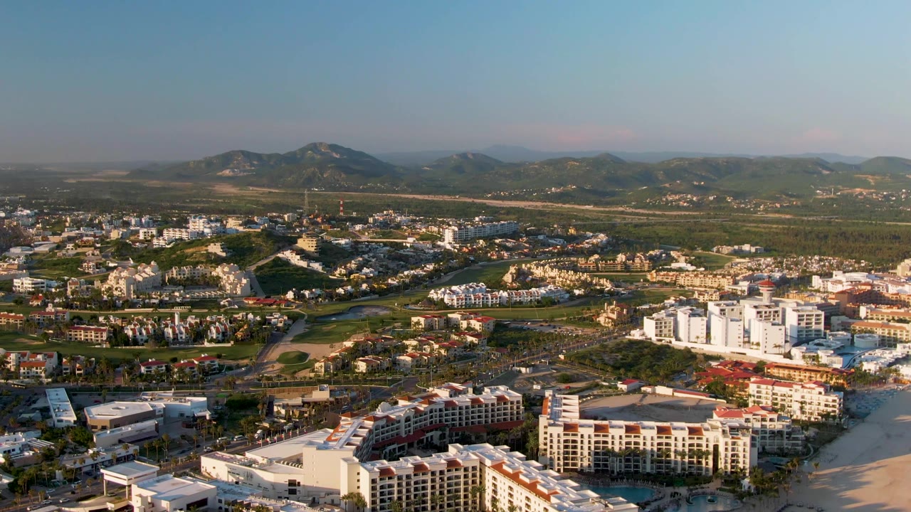 Wide drone-style view of resort buildings, beach, roads, greenery, and hazy mountains.