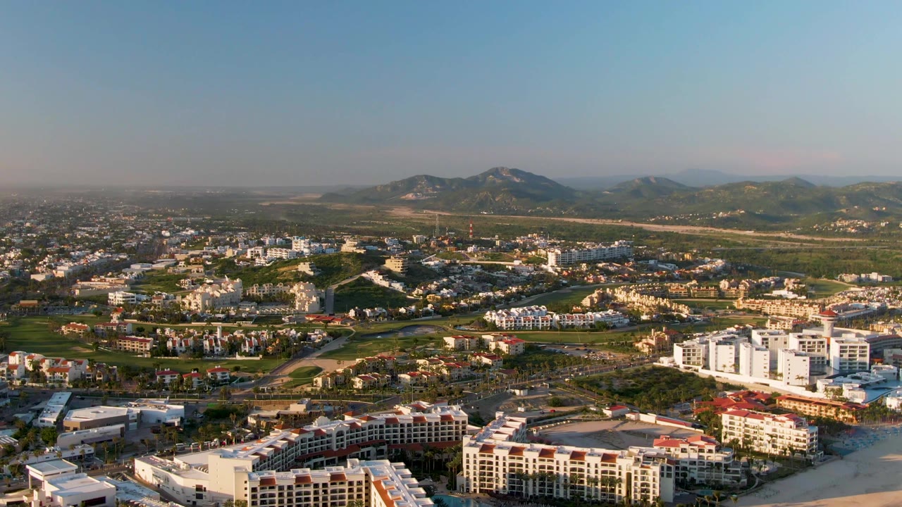 San Jose del Cabo appears from a high aerial angle with a sandy shoreline at the bottom edge, dense low-rise development, green areas, distant hills, and no visible people.