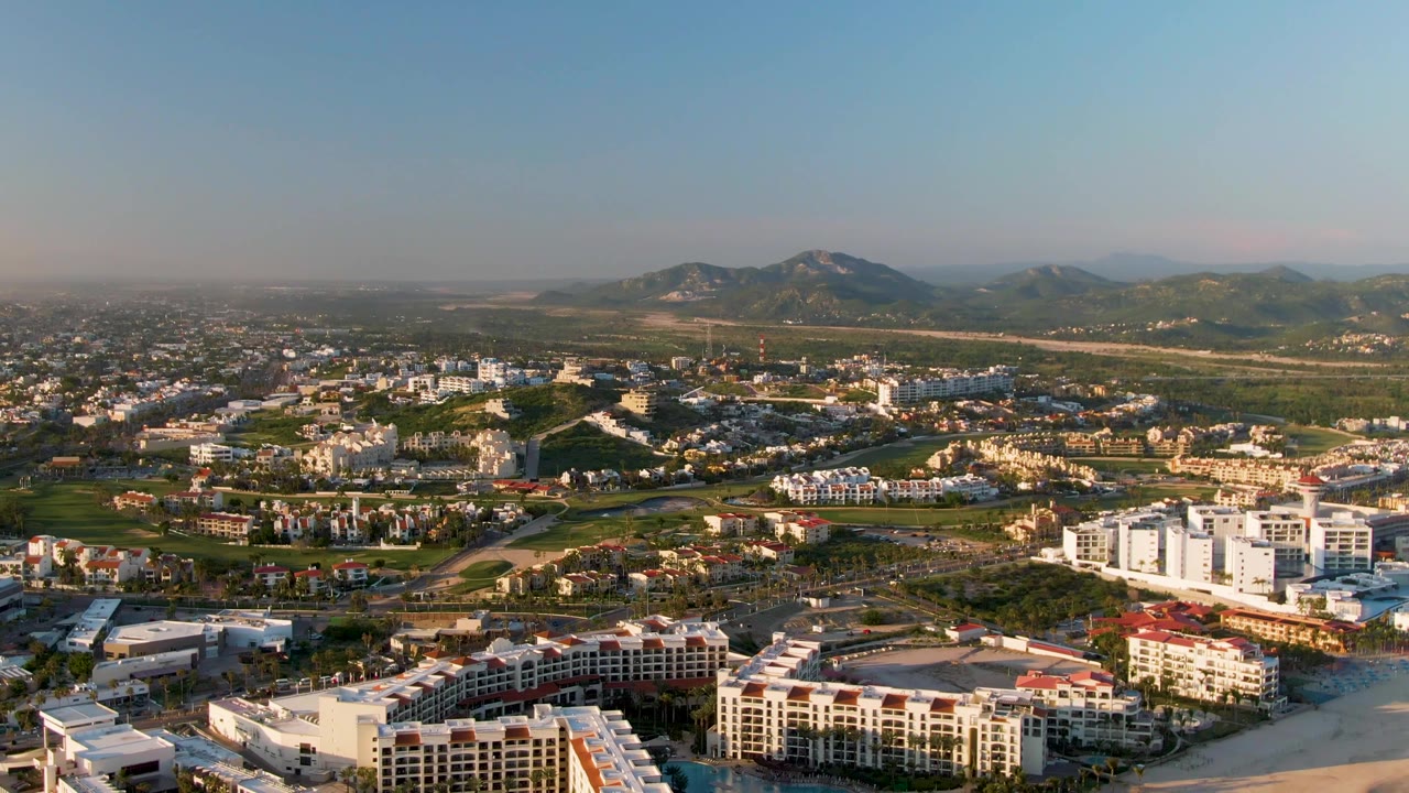 Mid-clip aerial frame over San Jose del Cabo showing bright beachfront structures, a spread of town buildings, green terrain, distant mountains, and no visible people.
