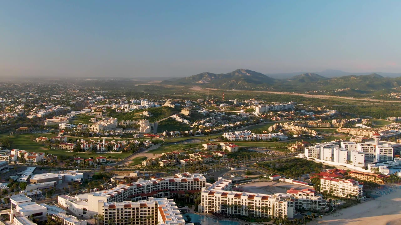 A high aerial view of San Jose del Cabo shows resort-style buildings near the beach, inland neighborhoods, patches of greenery, muted haze near the horizon, and no visible people.