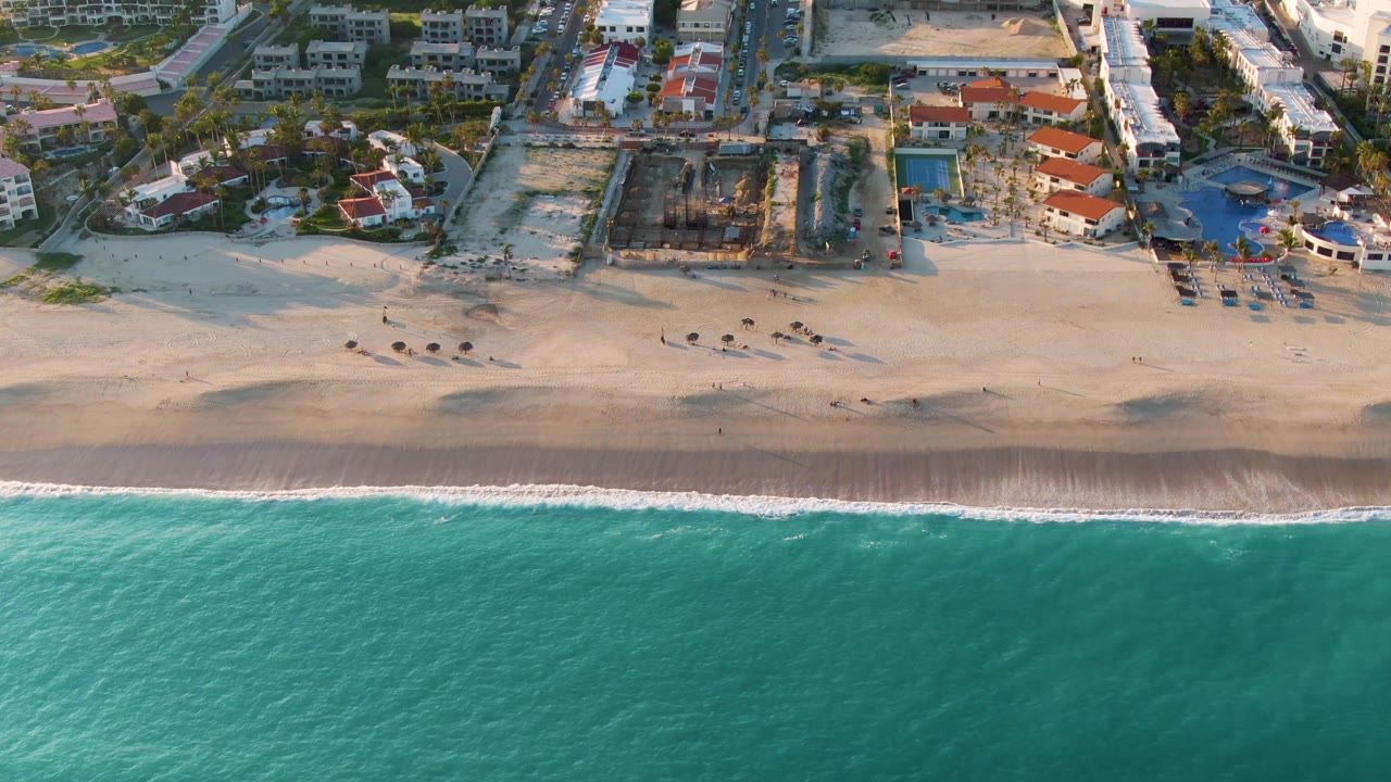 Aerial frame from San Jose Del Cabo showing turquoise water below a narrow surf line, wet and dry sand, and beachfront buildings and pools with no recognizable people visible.