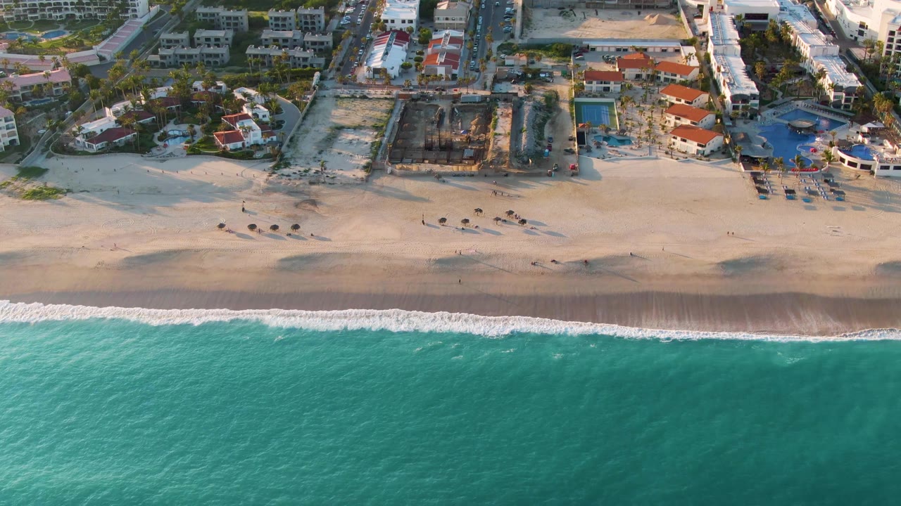 Aerial beach frame with waves reaching the sand near pools and low buildings.