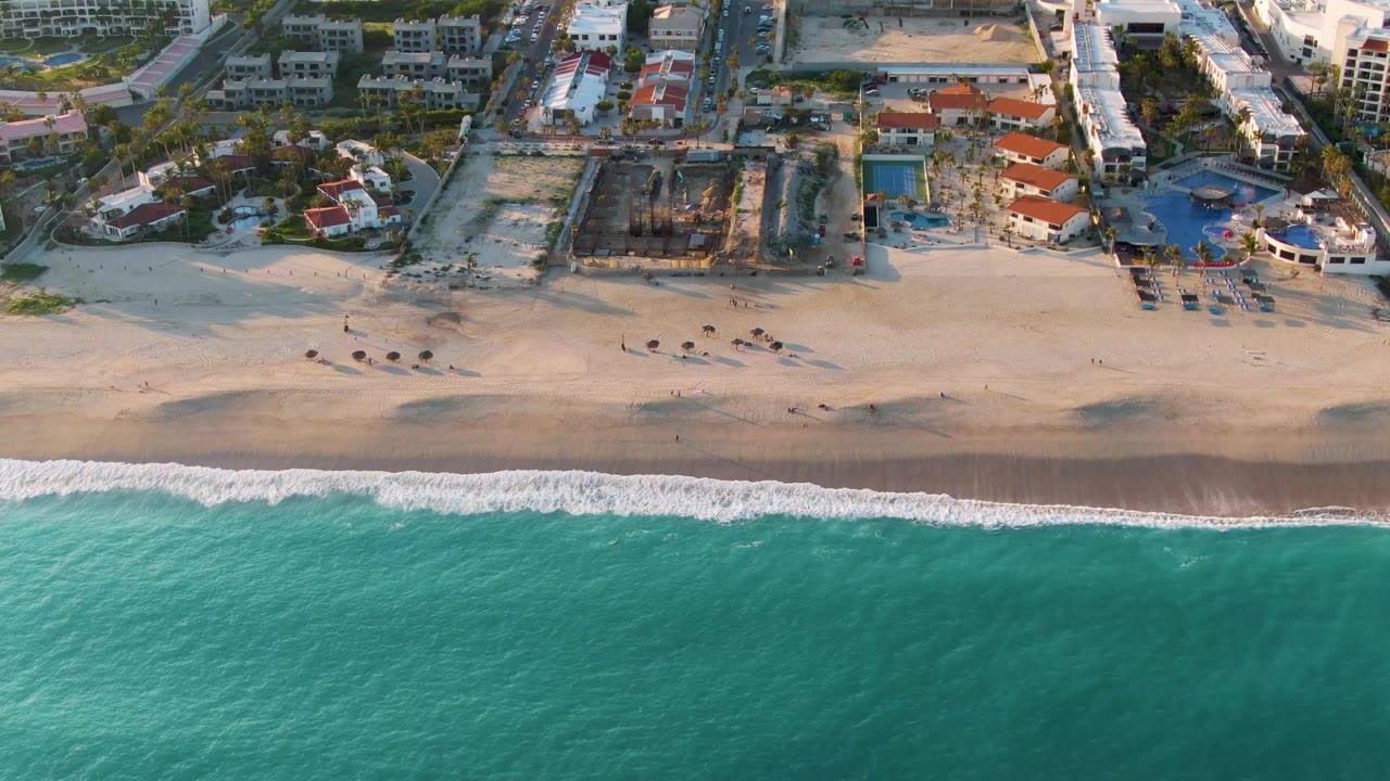 Aerial San Jose Del Cabo frame with teal water occupying the lower half, a clean white wave band at the shoreline, and a cluster of pools and buildings casting shadows inland.
