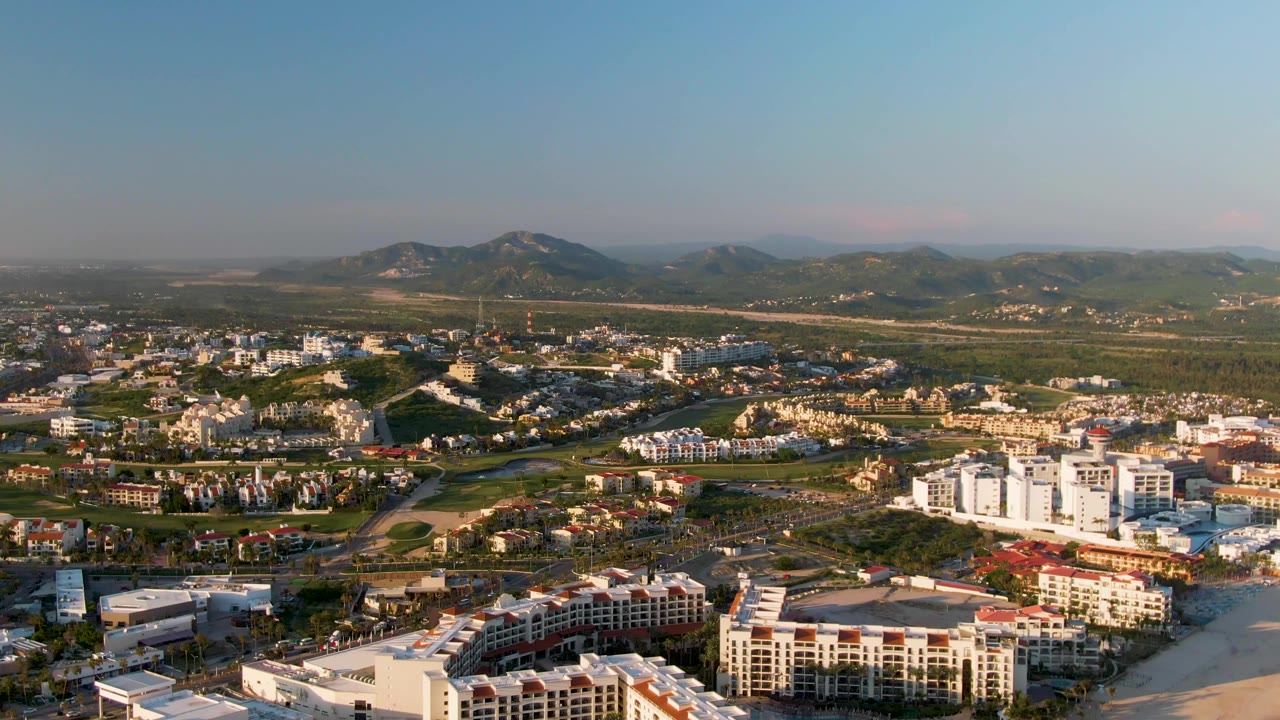 Wide aerial view of San Jose Del Cabo with sandy beach buildings in the foreground, urban development across the middle distance, mountains on the horizon, and no visible people.