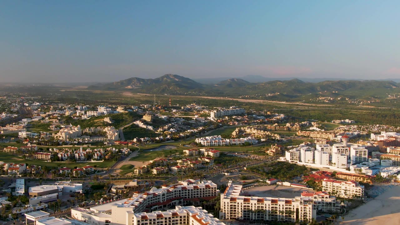 Aerial frame from San Jose Del Cabo showing coastal buildings at the bottom edge, a spread of roads and green open land in the midground, hazy mountains beyond, and no visible people.