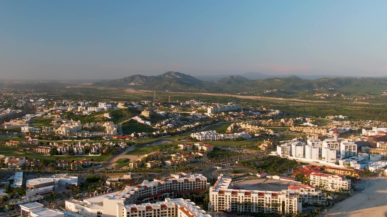 San Jose Del Cabo aerial still with white mid-rise buildings near the beach, lower residential and commercial blocks behind them, distant mountains, clear sky, and no visible people.